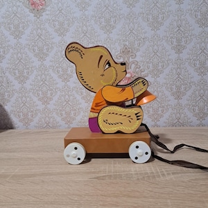 Puede incluir: Un juguete de madera vintage para tirar que representa un oso de dibujos animados. El oso, vestido con ropa naranja y morada, está sentado en un carro de madera marrón con ruedas blancas. Una cuerda marrón está unida.