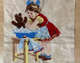 Peut inclure: Une tapisserie vintage représentant une jeune fille lavant un ours en peluche dans une bassine bleue. La fille porte une robe rouge, un haut jaune et une jupe bleue. La scène se déroule sur un petit tabouret en bois, le tout rendu en fil coloré.