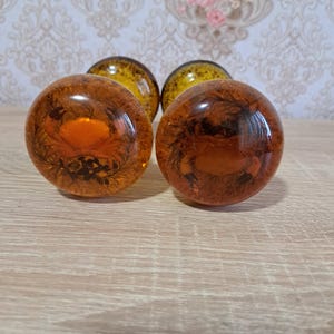 Peut inclure: Quatre boutons de meuble ronds, de couleur ambre, avec un motif décoratif organique à l'intérieur. Les boutons sont disposés en rangée sur une surface en bois clair, avec un fond d'écran floral.