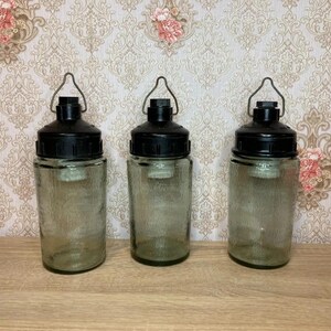 May include: Three glass lanterns with black tops and metal handles. The lanterns have a textured surface and are cylindrical in shape. They are displayed on a wooden surface against a patterned wallpaper background. The lanterns are decorative.