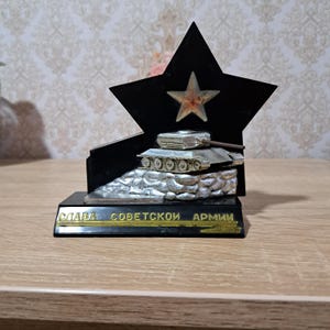 Peut inclure: Trophée noir en forme d'étoile avec un modèle de char d'assaut argenté sur une base rocheuse. Le trophée comporte une étoile dorée au centre et le texte "Gloire à l'Armée soviétique" en lettres dorées.