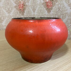 May include: A round, red ceramic pot with a dark rim and base. The pot has a bulbous shape and sits on a light-colored wooden surface. The background features a patterned wallpaper.
