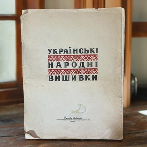 Op de afbeelding: Een vintage boekomslag met de titel "Українські народні вишивки" in zwarte tekst op een witte achtergrond. De titel is omlijst door een rood en zwart geborduurd ontwerp.