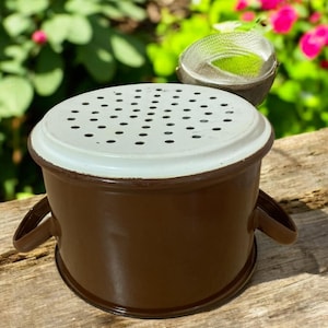 Puede incluir: Una olla de esmalte marrón con una tapa blanca perforada. La olla tiene dos asas marrones y un borde marrón. Una pequeña cesta de malla es visible en el fondo, con un fondo floral verde y rosa borroso.