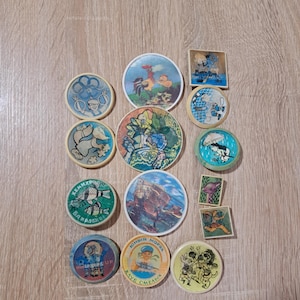May include: A collection of vintage circular and square collectible pins with colorful illustrations. The pins feature various cartoon characters, animals, and scenes, with text in Cyrillic script. The pins are arranged on a light brown wooden surface.