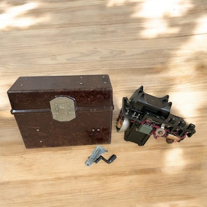 May include: A vintage brown rectangular box with a metal clasp and a disassembled black device with wires and gears. A small metal crank is also present, all on a wooden surface.