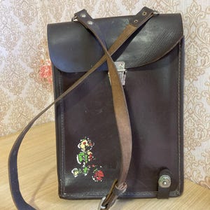 May include: A dark brown leather satchel with a flap closure and a long shoulder strap. The satchel features a colorful cartoon graphic on the front. The bag has a metal clasp and a decorative button on the bottom.
