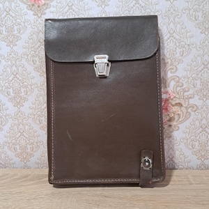 May include: A brown leather portfolio with a flap closure and a silver-colored clasp. The portfolio has a rectangular shape and a small strap with a button closure on the bottom. The stitching is visible along the edges.