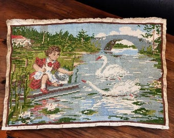 May include: A vintage needlepoint tapestry depicting a serene waterscape. The scene features a young person in a red dress, two swans, a bridge, and a house. The tapestry has a fringed border and is set against a wooden surface.