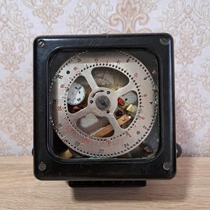 Könnte beinhalten: Ein schwarzer Vintage-Mechanik-Timer mit einem transparenten Zifferblatt, das interne Zahnräder und ein kreisförmiges Zifferblatt mit Zahlen zeigt. Der Timer ist in einem quadratischen Rahmen eingeschlossen.