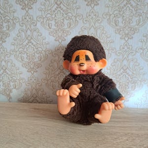 Może przedstawiać: Zabytkowa pluszowa lalka małpki z brązowym futrem, pomarańczową twarzą i czarnymi oczami. Lalka siedzi z wyciągniętymi ramionami i nogami. Tło przedstawia wzorzystą tapetę i drewnianą powierzchnię.