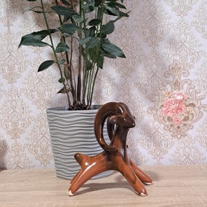 May include: A brown ceramic ram figurine with curved horns and white accents on its hooves. The figurine is displayed on a wooden surface, with a potted plant and patterned wallpaper in the background. The ram is a decorative item.