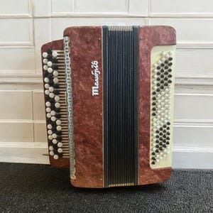 May include: A maroon accordion with black and white keys. The accordion has a textured finish and the text "Мелодия" in white script. The bellows are black, and the keys are arranged in vertical rows.