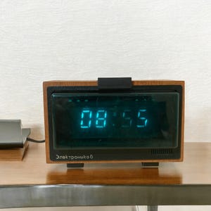 May include: A vintage digital clock with a wooden frame displays the time 08:55 in bright blue digits. The clock has a black top piece and the text "Электроника 6" is visible below the display. The clock is set on a wooden surface.