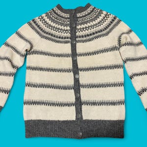 Könnte beinhalten: Eine cremefarbene, graue und anthrazitfarbene Strickjacke mit Knöpfen. Der Pullover hat horizontale Streifen und einen gemusterten Einsatz um den Kragen. Die Bündchen und der Saum sind anthrazitfarben. Der Pullover hat einen hohen Kragen und wird vorne mit Knöpfen geschlossen.