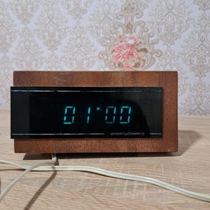 May include: A vintage digital clock with a wooden frame displays the time as 01:00 in blue digits. The clock has a rectangular black face and a light-colored power cord.