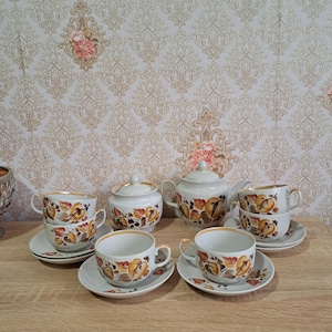 May include: A vintage tea set featuring a teapot, sugar bowl, and six teacups with saucers. The white porcelain set is decorated with a floral pattern in shades of orange, yellow, and brown, with gold trim. The set is arranged on a wooden surface.