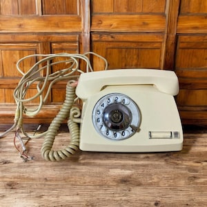 Puede incluir: Teléfono de disco rotatorio vintage de color crema con un cable de auricular en espiral y un dial con números. El teléfono está sobre una superficie de madera con un fondo de madera. El teléfono tiene forma rectangular con la parte superior curvada.