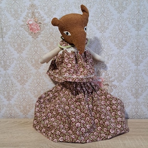 Könnte beinhalten: Eine handgefertigte Plüschpuppe mit braunem Kopf und langer Schnauze, die ein Kleid mit Blumenmuster trägt. Das Kleid ist braun mit kleinen rosa und weißen Blumen. Die Puppe hat kleine Arme und einen grünen Kragen. Der Hintergrund ist eine Blumentapete.
