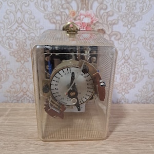 May include: A vintage electrical timer encased in a clear, rectangular box. The timer features a circular gauge with a black needle and numerical markings. Metal components are visible within the case, which is secured by a metal clasp.
