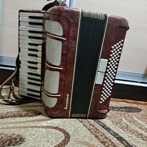 May include: A burgundy accordion with a black and white keyboard and a series of white buttons. The accordion has a decorative design on the front and a shoulder strap. The word "Sonuma" is written on the front.