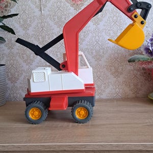 May include: A red, white, and yellow toy excavator. The excavator has a red arm and bucket, a white cab, and black wheels with yellow centers. The toy is designed for children and is made of plastic.