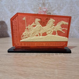 以下が含まれることがあります： 黒いベースが付いた、長方形の赤オレンジ色の装飾品。前面には、馬に引かれた戦車のクリーム色のレリーフがあります。テキストは、部品の側面と底面に表示されます。