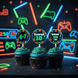 May include: Three cupcakes with teal frosting and colorful sprinkles, each topped with a round topper. The toppers feature gaming-themed designs and text that reads "6-7", "COOPER 10", and "BRUH DIG IN!". The background has neon gaming graphics.