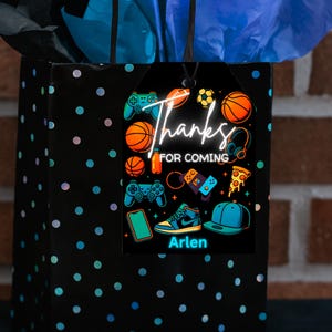 May include: A black gift bag with blue and pink polka dots, topped with blue tissue paper. A black tag features gaming and sports graphics, the words "Thanks for Coming," and the name "Arlen."
