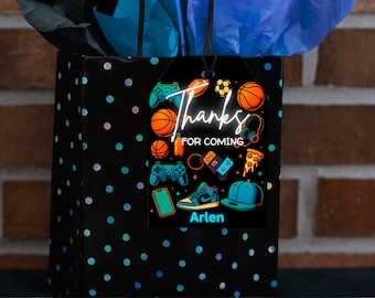 Può includere: Un sacchetto regalo nero con pois blu e rosa, sormontato da carta velina blu. Un'etichetta nera presenta grafica di giochi e sport, le parole "Thanks for Coming" e il nome "Arlen."