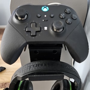 May include: A black Xbox controller with a green light on the front is mounted on a wall. A black Turtle Beach headset with green accents is also mounted on the wall.