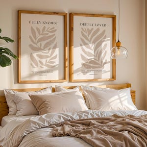 May include: Two framed prints with beige leaf illustrations and inspirational quotes, "Fully Known" and "Deeply Loved", hanging above a bed with white bedding and a beige throw blanket.