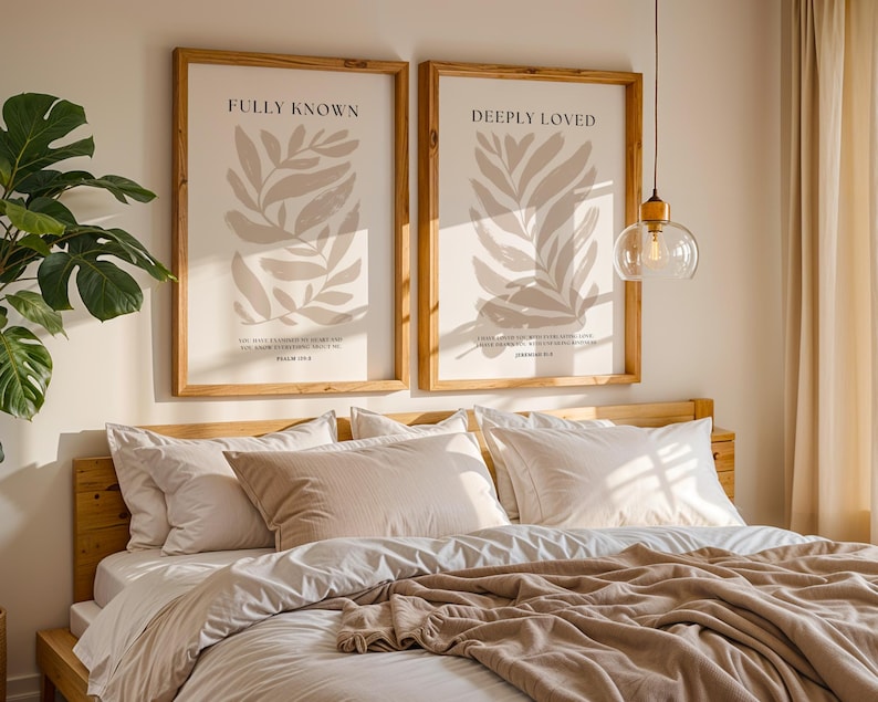 May include: Two framed prints with beige leaf illustrations and inspirational quotes, "Fully Known" and "Deeply Loved", hanging above a bed with white bedding and a beige throw blanket.