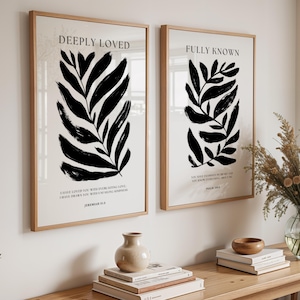 May include: Two framed art prints with botanical designs. One print reads "DEEPLY LOVED" and the other reads "FULLY KNOWN". Both prints feature black leaf-like shapes on a cream background. The frames are light wood.