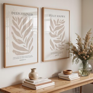 May include: Two framed prints with beige leaf illustrations and inspirational quotes. The left print says "Deeply Loved" and the right print says "Fully Known". The prints are hanging on a white wall above a wooden table with books, a vase of dried flowers, and a wooden bowl.