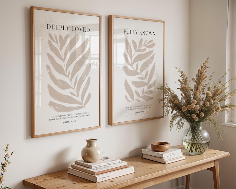 May include: Two framed prints with beige leaf illustrations and inspirational quotes. The left print says "Deeply Loved" and the right print says "Fully Known". The prints are hanging on a white wall above a wooden table with books, a vase of dried flowers, and a wooden bowl.