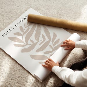 May include: A person unrolling a white poster with a brown abstract leaf design and the text "FULLY KNOWN" printed on it.