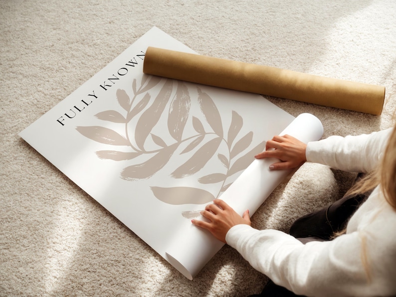 May include: A person unrolling a white poster with a brown abstract leaf design and the text "FULLY KNOWN" printed on it.
