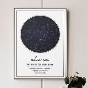 Op de afbeelding: Ingelijste print met een donkerblauwe cirkelvormige sterrenkaart. De print bevat de naam "olivia" in cursief, de tekst "THE NIGHT YOU WERE BORN" en details over locatie en datum. De lijst is wit.