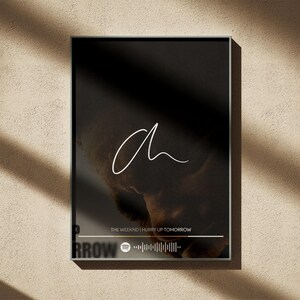Puede incluir: Impresi&oacute;n art&iacute;stica enmarcada con fondo negro, una "a" blanca en cursiva y una imagen borrosa. El texto "THE WEEKND | HURRY UP TOMORROW" se muestra debajo, junto con un c&oacute;digo de Spotify. Marco oscuro.