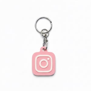 Könnte beinhalten: Ein rosa Schlüsselanhänger mit weißem Instagram-Logo. Der Schlüsselanhänger ist quadratisch mit abgerundeten Ecken und einem silbernen Metallring und -verschluss. Das Instagram-Logo ist eine weiße Umrandung einer Kamera.