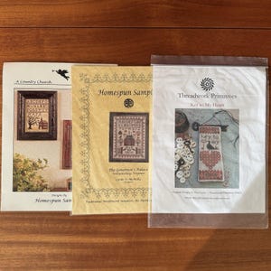 May include: Three rectangular paper patterns for cross-stitch embroidery. The left shows A Country Church, the center shows Homespun Sampler The Governor's Palace, and the right shows Threadwork Primitives Key to My Heart with a bird and key motif.