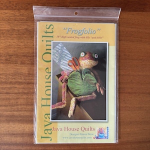 May include: A Java House Quilts sewing pattern package titled Frogfolio, featuring a 10-inch high seated frog with a lilly pad-folio. The packaging displays a photograph of a fabric frog holding orange scissors and a green spool of thread.