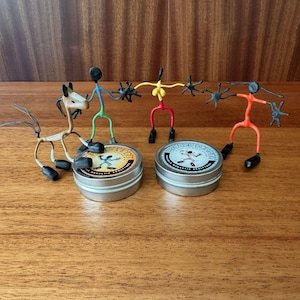 May include: Three colorful wire stick figures with black magnetic hands and feet stand near two round silver metal tins. One figure is yellow, one is orange, and one is blue and green. A tan wire horse figure stands beside the tins labeled DENDY DANGER.