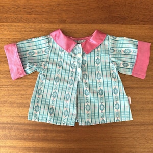 May include: A doll's shirt with a blue and white plaid pattern, featuring pink collar and cuffs. The shirt has a button-up front and the letters "CPK" are printed throughout the design.