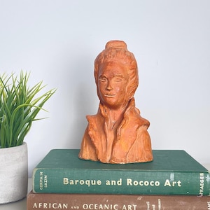 Peut inclure: Un buste en terre cuite d'une personne avec un chignon, posé sur deux livres. Le livre du dessus est vert et dit "Baroque and Rococo Art". Le livre du dessous est brun et dit "African and Oceanic Art".