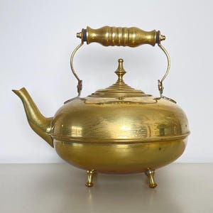Puede incluir: Una tetera de latón vintage con asa de madera y tres patas. La tetera tiene un cuerpo redondo y una tapa abovedada.