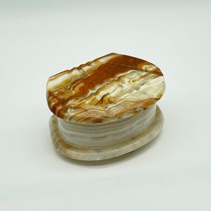 May include: A small, oval-shaped box made of white and brown onyx. The top of the box is carved with a swirling pattern.