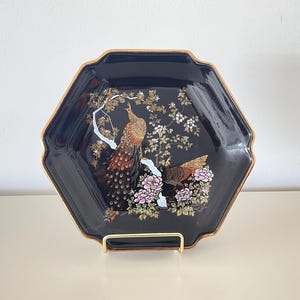 Può includere: Un piatto decorativo nero, esagonale, con bordo dorato. Il piatto presenta una dettagliata illustrazione di due pavoni appollaiati su un ramo fiorito. I pavoni hanno piume marroni e dorate, e i fiori sono rosa e bianchi.