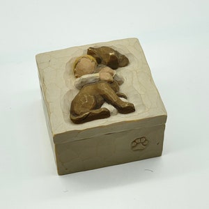 Puede incluir: Caja rectangular beige con un relieve tallado de un niño abrazando a un perro. La tapa presenta al niño y al perro, y una huella de pata está en el lateral. La caja está hecha de un material claro.